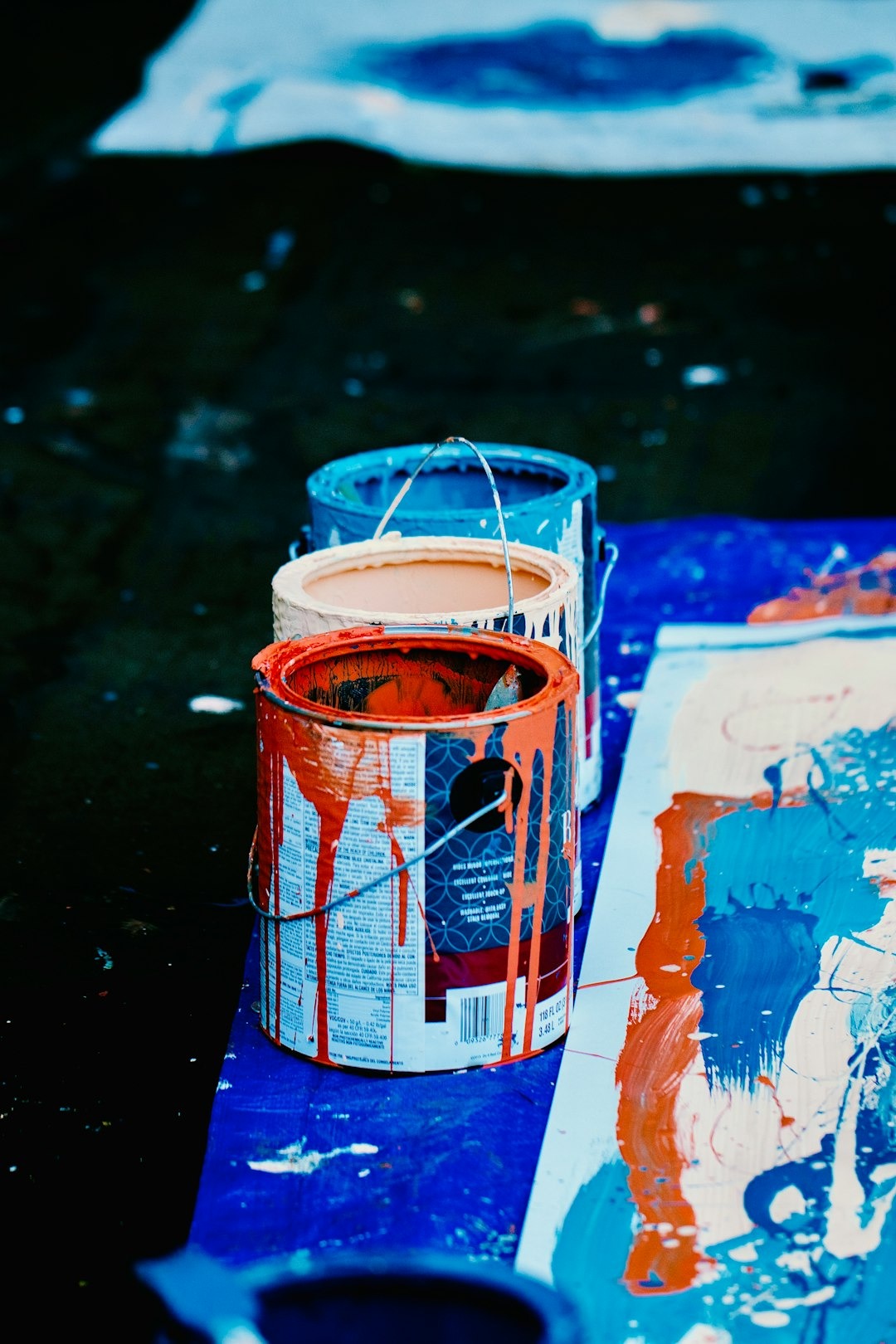 Three open paint cans with drips lined up on a blue tarp.