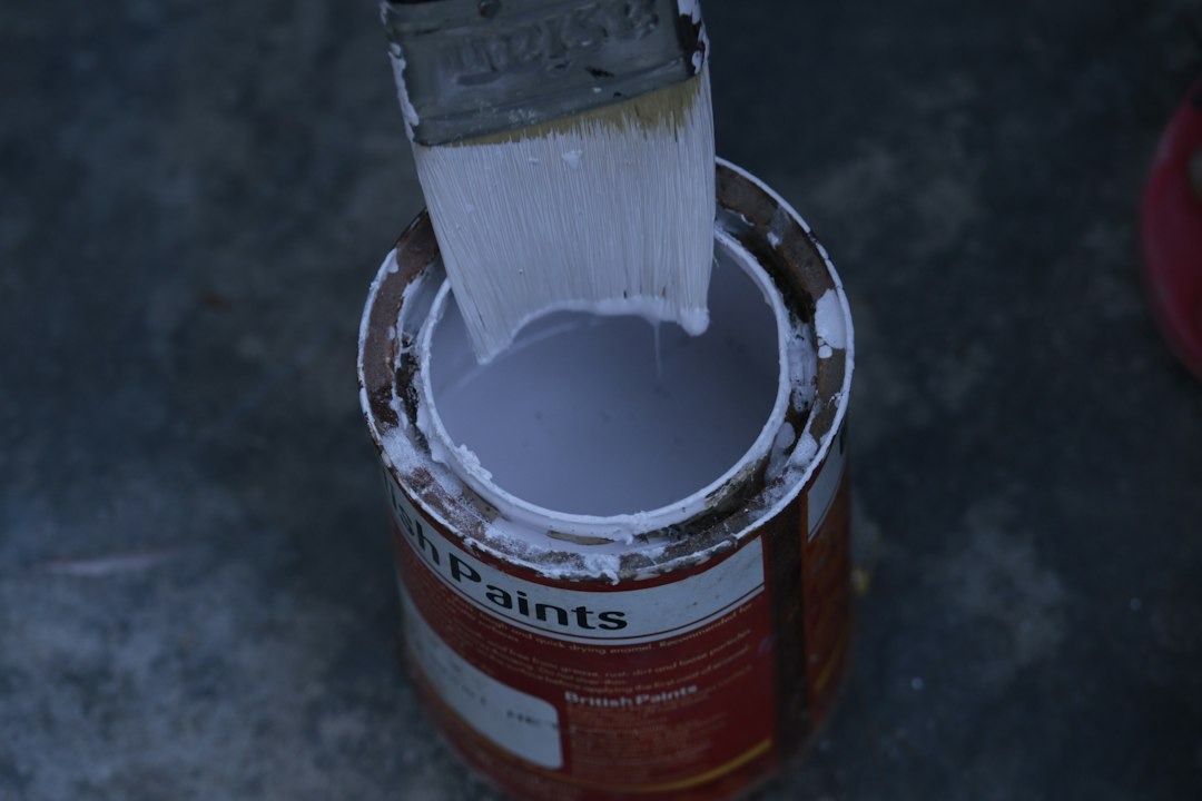 A paintbrush drips white paint into a worn red can.